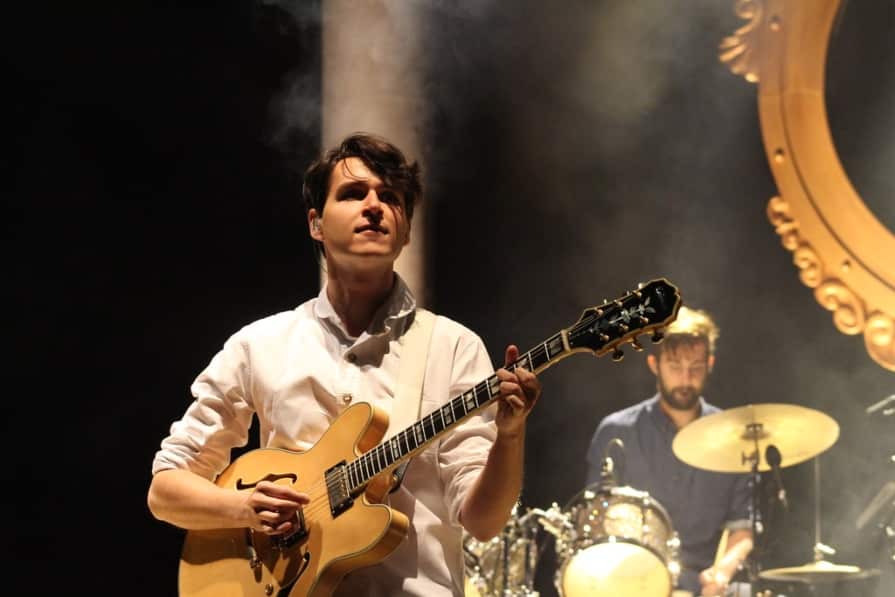 A guitarist in a white shirt passionately performs on stage with a drummer in the background amidst theatrical stage lighting and a hazy atmosphere, creating an intimate Vampire Weekend live music scene.